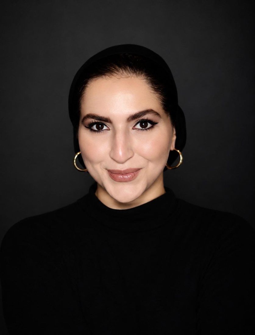Naeema Hasan in a black turtleneck and headscarf with gold hoop earrings, smiling