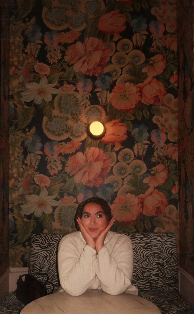 Naeema smiling with her hands beneath her chin, seated under floral wallpaper and a glowing lamp