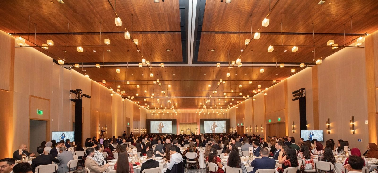 A warmly lit ballroom filled with guests at round tables, with a speaker at the podium shown on large screens